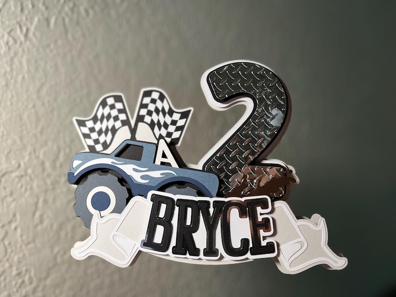 Może przedstawiać: Niebieski topper na tort w kształcie potwora z napisem "Bryce", z flagami w szachownicę i numerem 2.