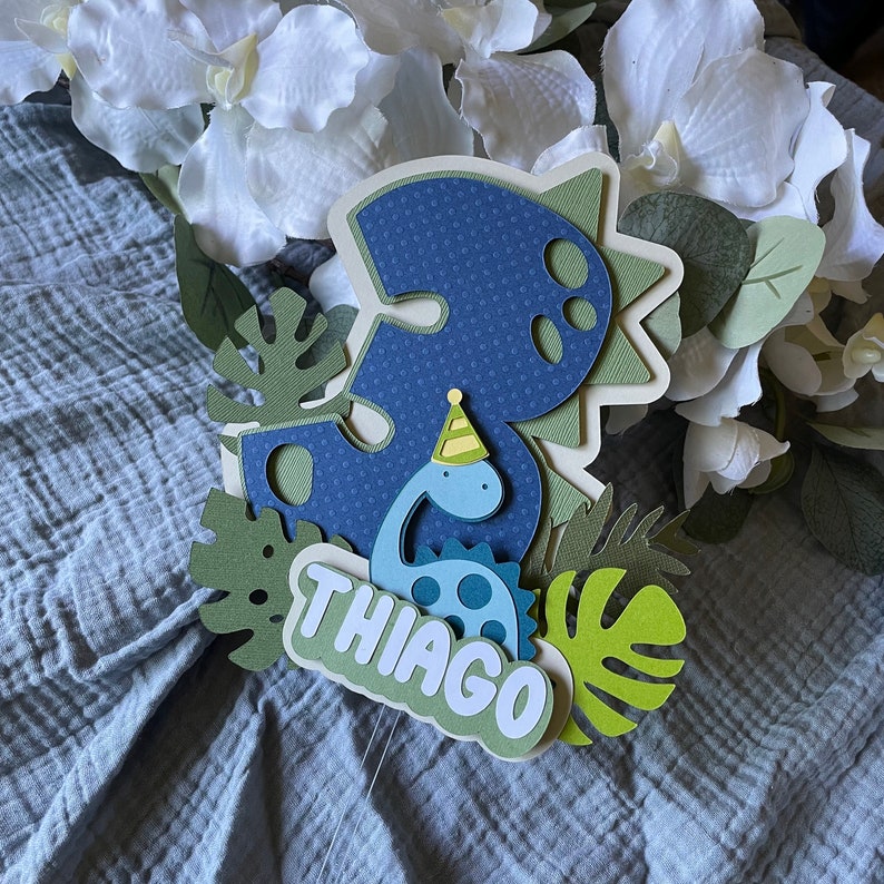 May include: A blue and green dinosaur cake topper with the number 3 and the name Thiago. The dinosaur is wearing a party hat.