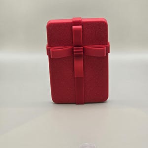 May include: A rectangular red jewelry box with a textured surface and a matching ribbon closure. The box is upright and centered, with the ribbon forming a cross on the front. The background is a neutral, light color.