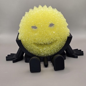 May include: A yellow sponge with eye cutouts and a smiling mouth, mounted on a black, stylized base with arms and legs. The sponge has a textured surface and is designed to resemble a cartoon character.