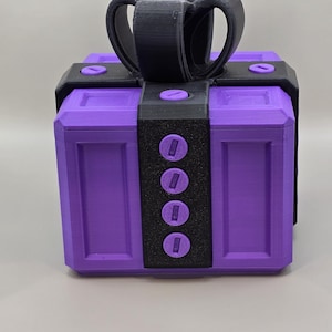 Puede incluir: Una caja morada con un lazo y detalles en negro. La caja tiene forma rectangular con una banda negra y cuatro detalles circulares en la parte delantera. El lazo está hecho de un material grueso y curvado.