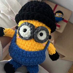 Op de afbeelding: Een gehaakt Minion-speelgoed met een zwarte hoed, een geel lichaam, een blauwe overall en grote witte ogen.