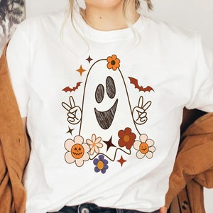 May include: White t-shirt with a cartoon ghost wearing flowers and making a peace sign. The ghost has a happy face and is surrounded by colorful flowers.