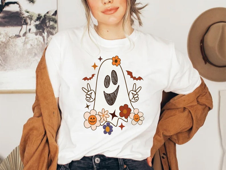 May include: White t-shirt with a cartoon ghost wearing flowers and making a peace sign. The ghost has a happy face and is surrounded by colorful flowers.