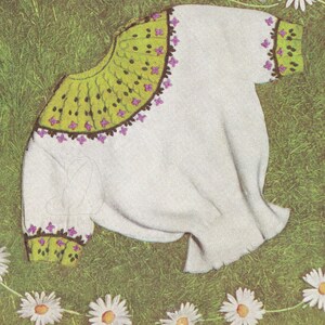 Könnte beinhalten: Ein weißer Strickpullover mit grünem und violettem Blumensaum an Ausschnitt und Ärmeln. Der Pullover liegt auf einem Bett aus grünem Gras.
