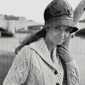 Puede incluir: Una foto en blanco y negro de una mujer con un cárdigan blanco de punto de cable con cierre de botón y un sombrero negro. El cárdigan tiene cuello y bolsillos.