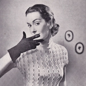 May include: A black and white photo of a woman wearing a white crocheted sweater with a cable knit pattern. She is wearing black gloves and a black belt.