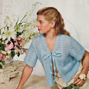 May include: A light blue short-sleeved cardigan featuring an open-front design with a matching satin ribbon tie closure at the neckline. The garment is crafted with a delicate, vertical lace-like knit pattern throughout the body and sleeves.