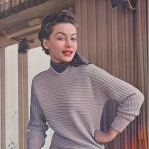 Puede incluir: Una mujer con un suéter gris claro y una bufanda de lunares negros y blancos. Está sosteniendo una revista en su mano izquierda y tiene la mano derecha en la cadera.