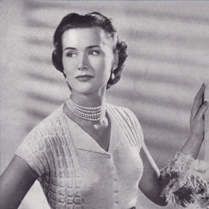May include: A black and white photo of a woman wearing a white knitted cardigan with a button closure and a pearl necklace. She is standing with her hand on her hip and looking to the side.