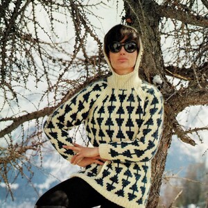 Puede incluir: Una mujer lleva un suéter blanco y azul con estampado geométrico de cuello alto y un cuello alto negro debajo. Lleva pantalones negros y botas negras de caña alta. Está de pie frente a un árbol con nieve en el suelo.