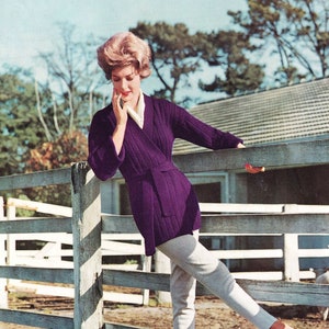 Könnte beinhalten: Eine Frau trägt einen lilafarbenen Strickcardigan mit Gürtel, weiße Hose und rote Schuhe. Sie steht vor einem weißen Holzzaun.