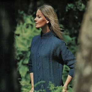 May include: A woman wearing a dark blue knitted sweater with a high collar and cable knit detailing. The sweater has long sleeves and is paired with a tan skirt. The image is set outdoors with a blurred green background.