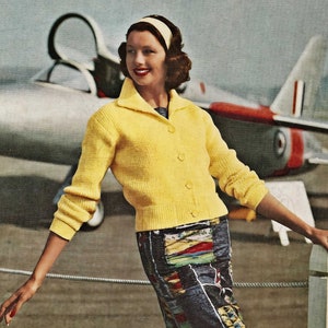 May include: A woman wearing a yellow cardigan with a button fastening and a colourful patterned skirt. She is standing in front of a white fence with a jet in the background.