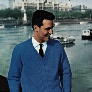 May include: A blue knitted sweater with a v-neck and cable knit detail. The sweater is worn by a person with a white shirt and a black tie.