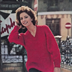 May include: A woman in a vibrant red V-neck sweater and a dark skirt. The sweater has a ribbed texture. The background shows a street scene with a building and a sign.