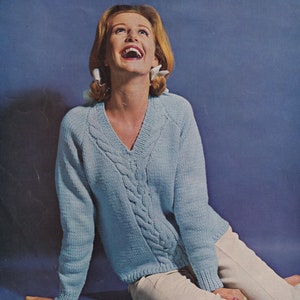 Puede incluir: Un suéter de punto azul claro con un patrón de trenza y cuello en V. El suéter lo lleva una mujer con cabello rubio y está sentada en una superficie azul.