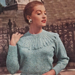 Op de afbeelding: Een lichtblauwe gebreide trui met een textuurontwerp en franjes rond de halslijn. De trui heeft lange mouwen en een geribbelde zoom. De vrouw op de foto draagt grote zilveren oorbellen.