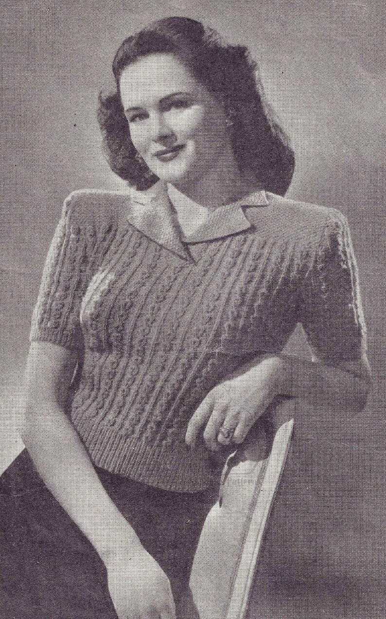 May include: A black and white photo of a woman wearing a cable knit sweater with a collared neckline. The sweater has a textured pattern and short sleeves.