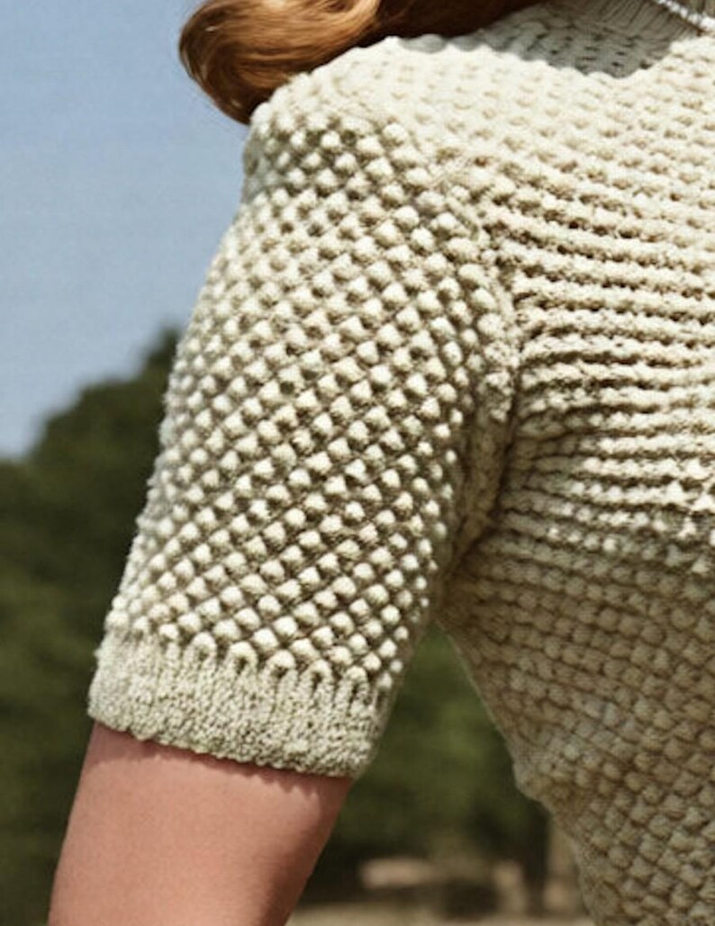 May include: A short-sleeved top in a light beige color featuring a prominent textured popcorn stitch pattern throughout the body and sleeves. The garment has a ribbed knit trim along the sleeve edge and a high neckline, showcasing a dense knit construction.