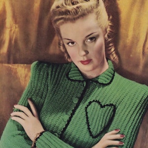 May include: A green knitted cardigan with a black heart-shaped detail on the front. The cardigan has a collar and buttons down the front.