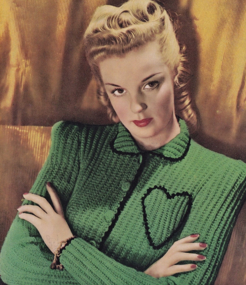 May include: A green knitted cardigan with a black heart-shaped detail on the front. The cardigan has a collar and buttons down the front.