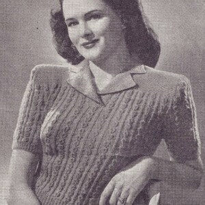 May include: A black and white photo of a woman wearing a knitted sweater with a cable knit pattern. The sweater has a collar and short sleeves.