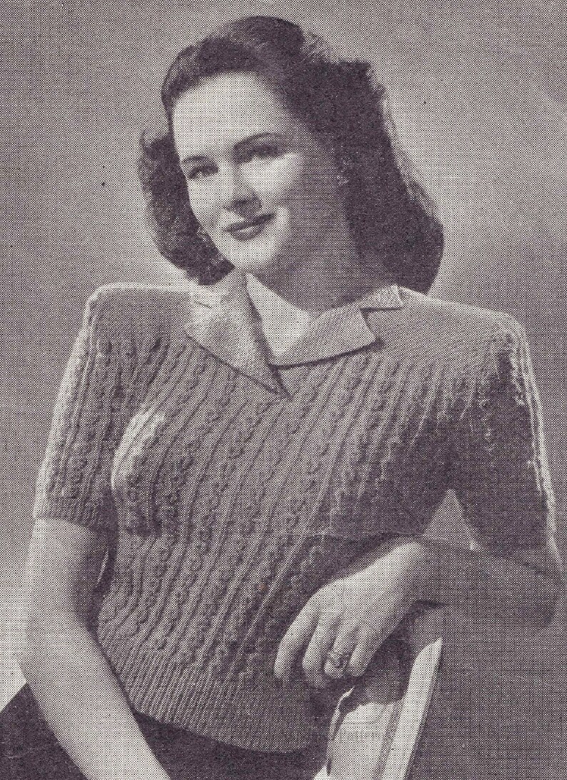 May include: A black and white photo of a woman wearing a knitted sweater with a cable knit pattern. The sweater has a collar and short sleeves.