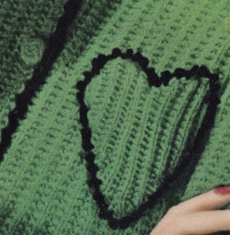 May include: A green knitted sweater with a black heart-shaped design. The sweater is made of a soft, textured yarn.