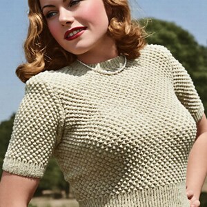 May include: A light green short-sleeved knit top featuring a textured popcorn stitch pattern, a ribbed crew neckline, and a wide ribbed waistband. The garment is worn with a simple white pearl necklace against a blurred outdoor background.