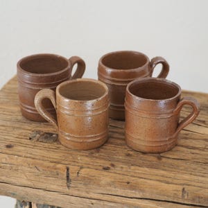 May include: A set of four small, cylindrical brown ceramic mugs with a speckled finish and two horizontal ridges encircling the body. Each mug features a single curved handle attached to the side, showcasing a consistent earthy tone and texture.