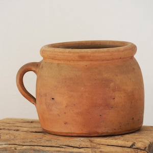 May include: A terracotta pot with a rounded body, a thick rim, and a single side handle. The surface of the vessel has a matte, earthy brown finish with subtle variations in color and texture, showing a simple, functional design.