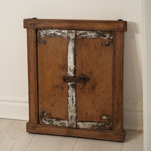 Peut inclure: Petit meuble ancien en bois avec un cadre rectangulaire. Les portes de l'armoire sont marron chaud avec des accents blancs et un loquet en métal. L'armoire est appuyée contre un mur blanc.