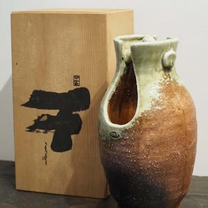 Puede incluir: Un jarrón de cerámica artesanal con un asa única y una superficie texturizada y moteada. El jarrón presenta un degradado de colores, pasando de una base marrón oscuro a una parte superior marrón claro y verde. Una caja de madera con caligrafía japonesa está al lado del jarrón.