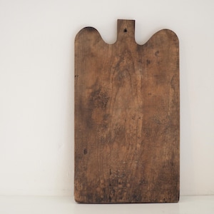 May include: A rectangular wooden cutting board with a weathered brown finish and visible grain patterns. The board features a rounded top edge with a central handle extension containing a small circular hole for hanging, showcasing a worn texture.