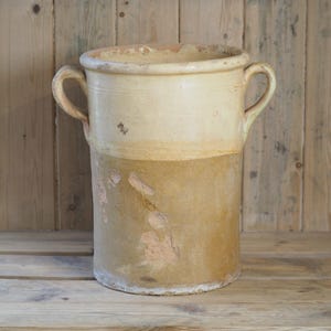 May include: A large, antique ceramic pot with two curved handles. The upper section is creamy white, the lower is a warm brown. The cylindrical pot shows signs of age and wear. It is likely used for storage or decorative purposes.