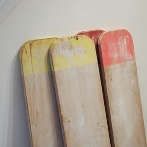 May include: Three wooden planks with rounded tops, painted with yellow and red accents. The planks are leaning against a white wall, showcasing their natural wood grain and distressed paint finish. The planks are likely for decorative purposes.