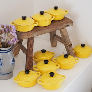 May include: A collection of eight small, round, bright yellow ceramic cocottes with matching lids and black knobs. The pots feature two side handles and a ribbed design. A grey ceramic pitcher with blue floral patterns holds purple dried flowers.