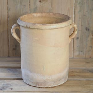 May include: A large, cylindrical, antique-style beige ceramic pot with two curved handles. The pot has a wide opening and a slightly textured surface. The pot is likely for decorative or functional use.