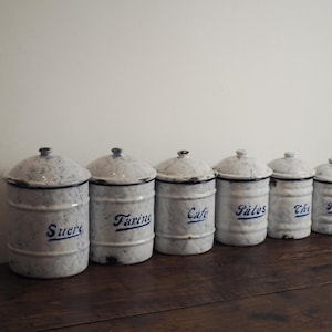 Puede incluir: Un conjunto de seis botes de esmalte blanco vintage con letras y tapas azules. Los botes están colocados sobre una superficie de madera oscura. Los botes tienen las palabras "Sucre", "Farine", "Cafe", "Sâles", "The" y "Poun".
