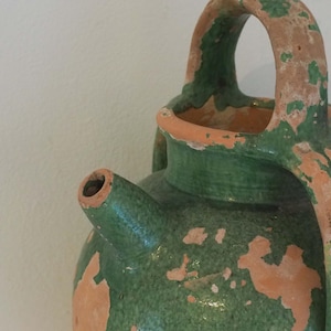 May include: A close-up of a rustic, green and orange ceramic jug. The jug has a spout, a large handle, and a smaller handle. The paint is distressed, giving it an aged appearance. The jug is likely used for decorative purposes.