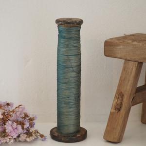 Op de afbeelding: Een hoge, cilindrische houten klos omwikkeld met dun, blauwgroen garen. De klos heeft donker, verweerd hout aan de boven- en onderkant, met tekenen van ouderdom en slijtage, en een gladde, verticale structuur die de vezel vasthoudt.