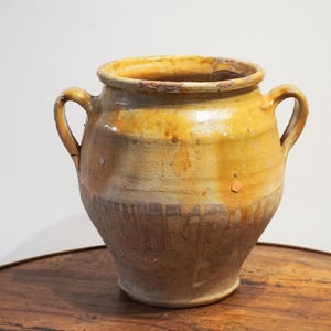 May include: A rustic, antique ceramic jar with two handles. The jar has a rounded body and a wide opening. It features a blend of yellow, brown, and beige tones, with visible signs of age and wear, suggesting it's an old piece.