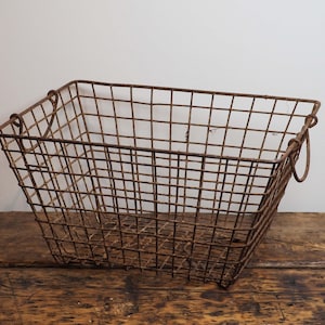 May include: A rectangular, rusty brown wire basket with rounded handles. The basket is made of a grid of metal wires, and sits on a wooden surface. The basket is empty.