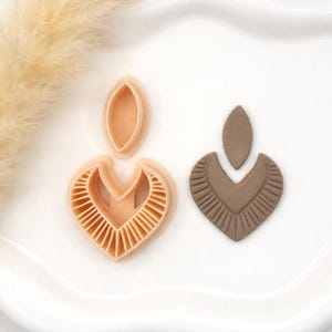 May include: A light peach-colored polymer clay earring cutter next to a finished brown clay earring. The cutter is in the shape of a heart with a teardrop top. The finished earring has a textured, fringed design.