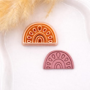May include: Two rainbow-shaped polymer clay cutters. One is a warm orange color with a white outline and the other is a dusty rose color with a similar white outline. The cutters are on a white surface.