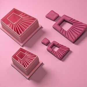 May include: Pink geometric earrings and matching box-shaped objects with a fan-like design. The earrings have a square top and a rectangular bottom with a cutout. The objects and earrings are on a pink surface.