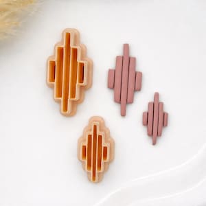 May include: Four geometric clay cutters and shapes in shades of peach and mauve. Two cutters are larger, with a diamond shape and vertical lines. Two smaller shapes are also diamond-shaped.