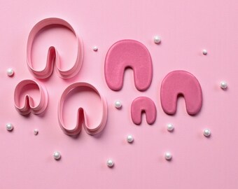 May include: A collection of pink horseshoe-shaped cookie cutters in various sizes, alongside several white pearl-like sprinkles, arranged on a solid pink background. The cutters are made of plastic and are designed for baking or crafting.