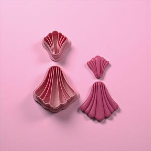 May include: Four pink seashell-shaped cookie cutters of varying sizes are arranged on a pink background. The cutters have a fluted design, with the largest measuring approximately 4 inches across. These tools are ideal for creating themed cookies or fondant decorations.